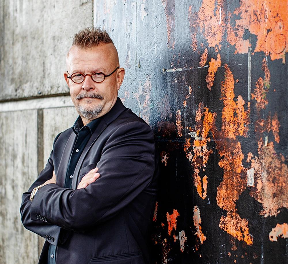 Man i svart kavaj och glasögon står med armarna i kors mot en vägg med flagnande färg och ser in i kameran.